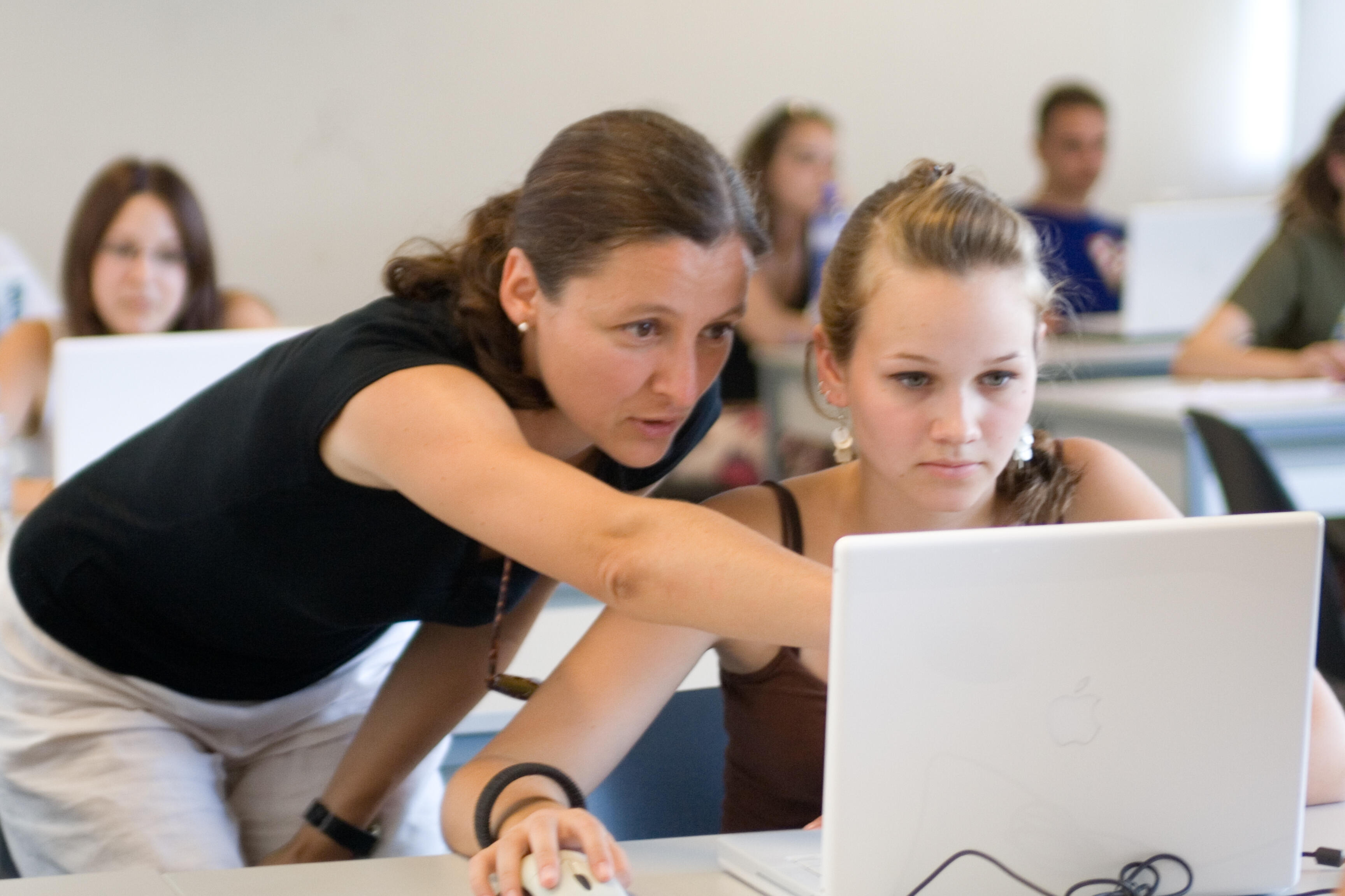 Eine Lehrerin zeigt einer Schülerin etwas auf einem Laptop-Bildschirm im Klassenzimmer.