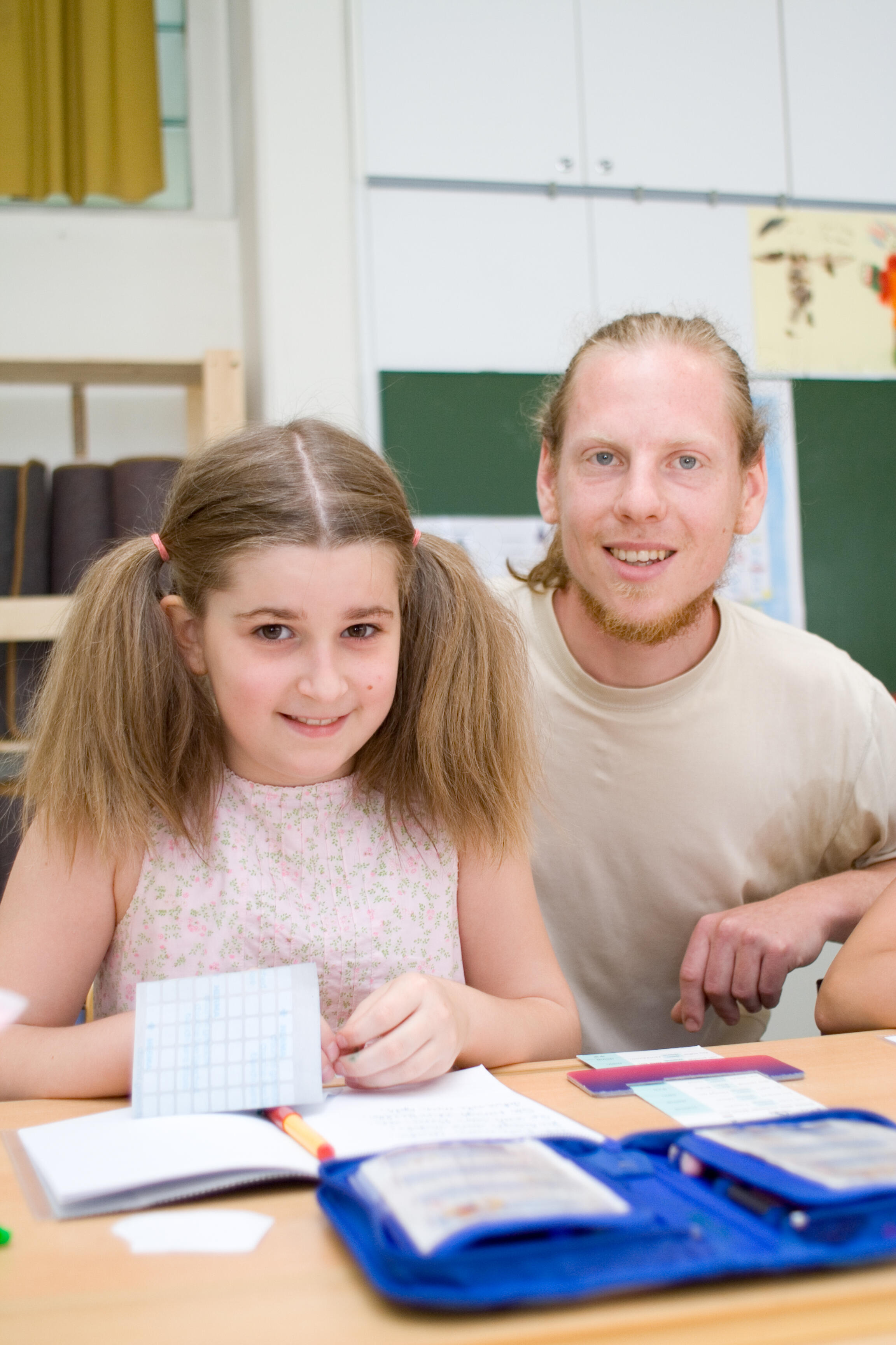 Junger Lehrer und Schülerin sitzen an einem Schreibtisch im Klassenraum, beide lächeln.