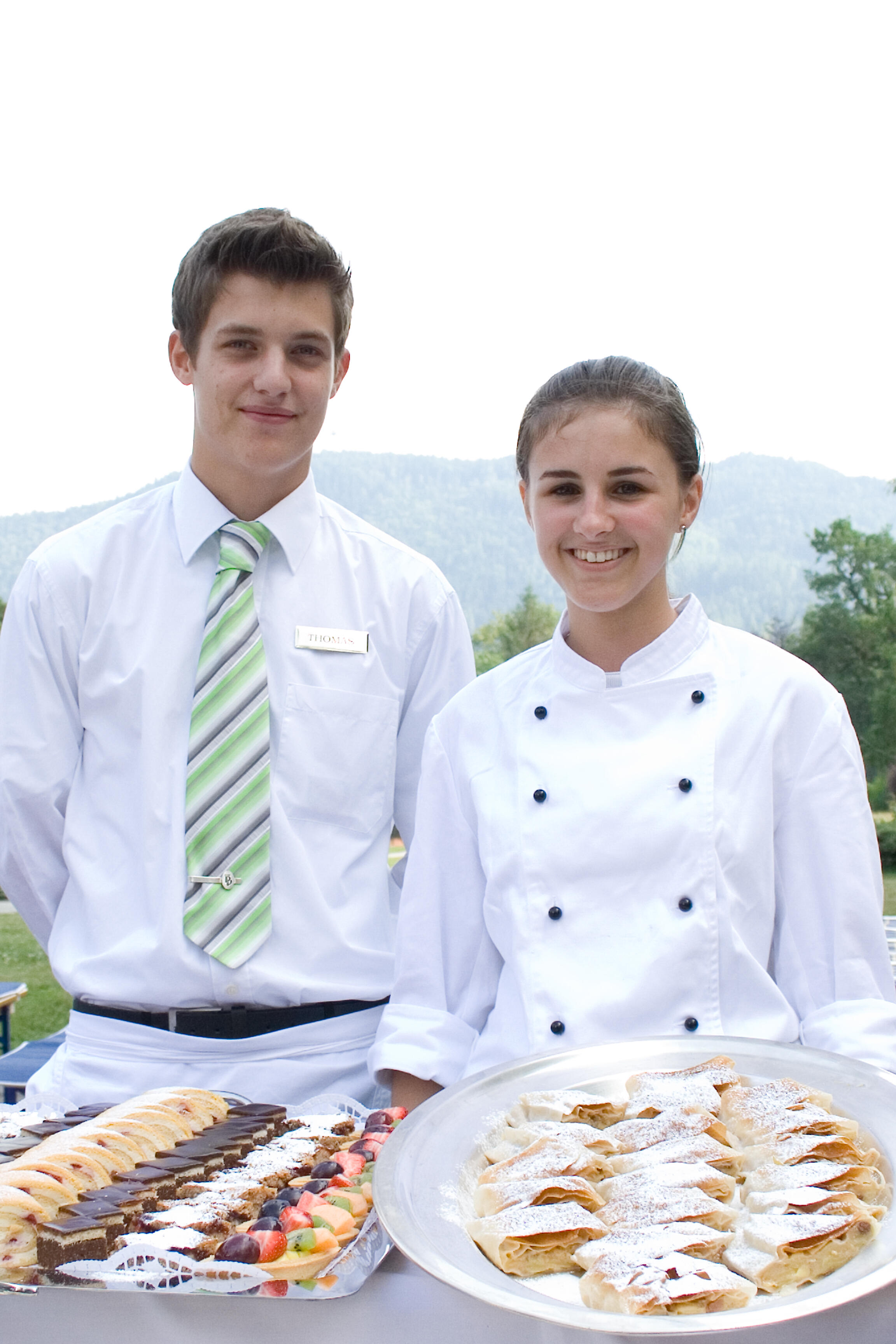 Zwei Lehrlinge, ein Bub und ein Mädchen in Gastronomie-Berufskleidung halten Tabletts mit Mehlspeien.
