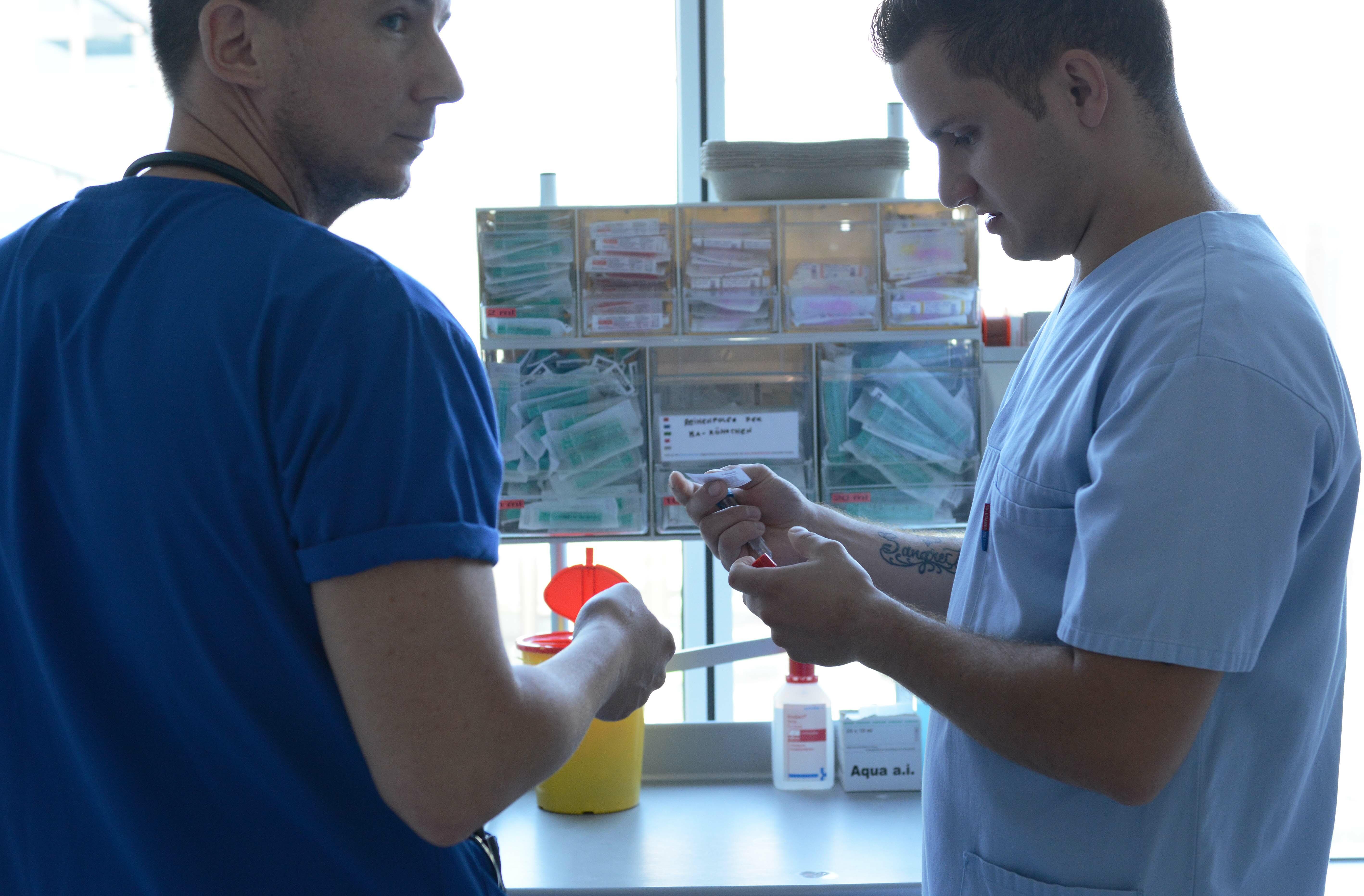 2 Pflegekräfte stehen vor einem durchsichtigen Aufbewahrungskästchen für Spritzen und Kanülen. Der jüngere Mann hält 2 Blutabnahmeröhrchen und ein Etikett in der Hand.