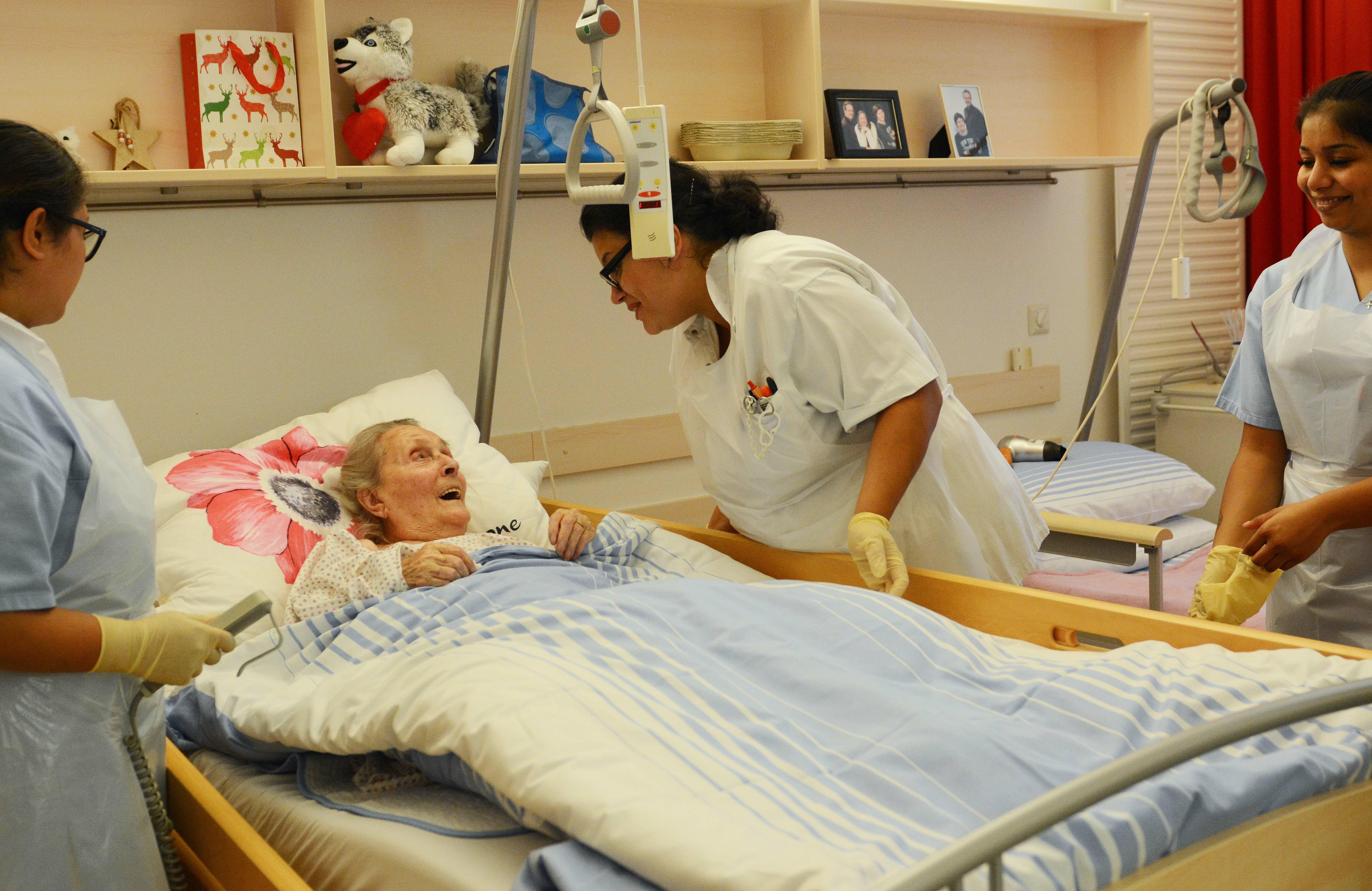 3 Pflegekräfte kümmern sich um eine alte Dame, die in einem Pflegebett liegt.