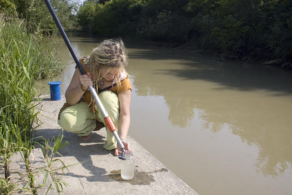 Eine junge Frau sitzt in der Hocke auf einem Steg an einem Gewässer und entnimmt eine Wasserprobe.