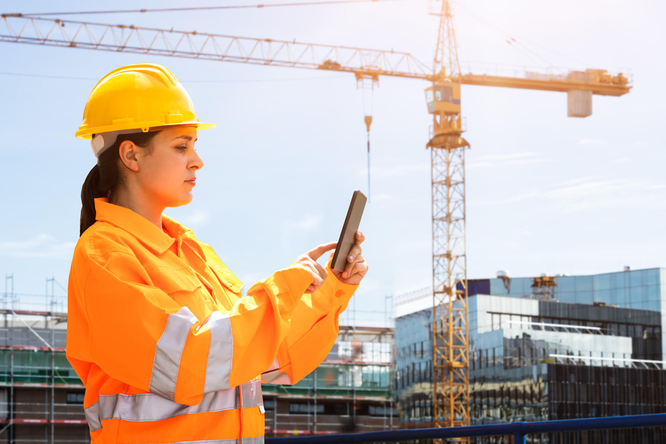 Eine Frau in Schutzkleidung und mit Schutzhelm bedient auf einer Baustelle ein Tablet. Im Hintergrund ist ein Kran zu sehen.