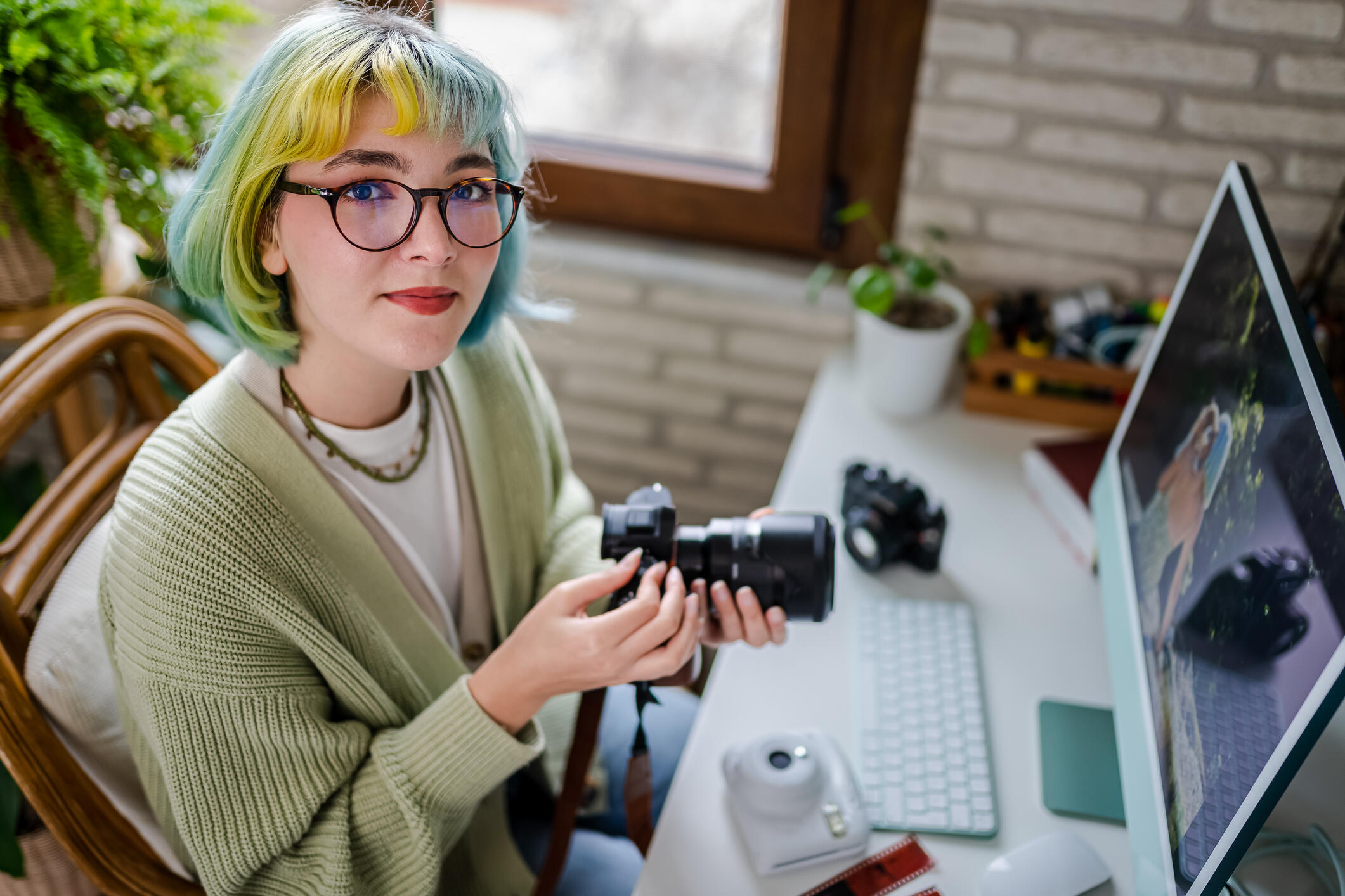 Junge Frau mit bunt gefärbtem Haar sitzt in Agentur an einem Schreibtisch und hat mehrere Fotokameras vor sich.