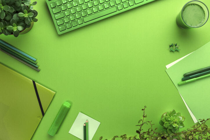 'Grün eingefärbtes Symbolfoto von einem Arbeitsplatz mit einer Tastatur, eine Mappe, Stifte, Papier, ein Getränk sowie Topfpflanzen.
