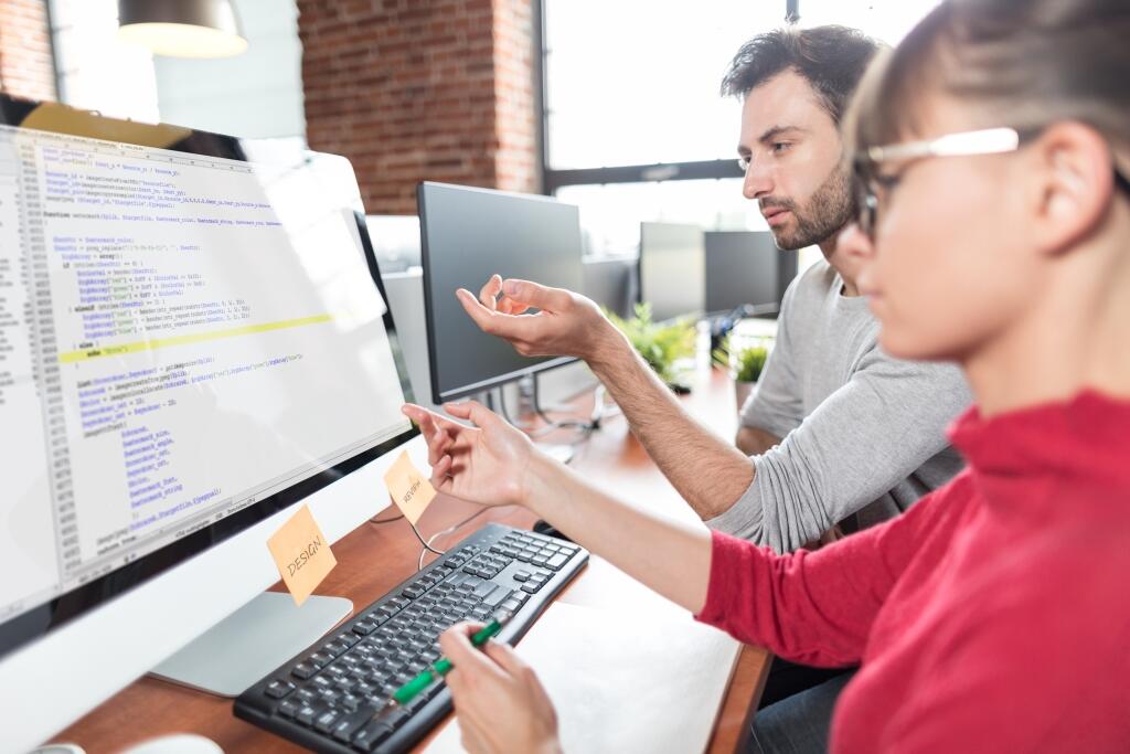 Eine Programmiererin und ein Programmierer sitzen an einem Computer-Arbeitsplatz. Auf dem Bildschirm vor ihnen sind Teile eines Codes zu sehen.