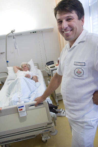 Ein lächelnder Pfleger steht am Fußende des Krankenbettes einer älteren Patientin.